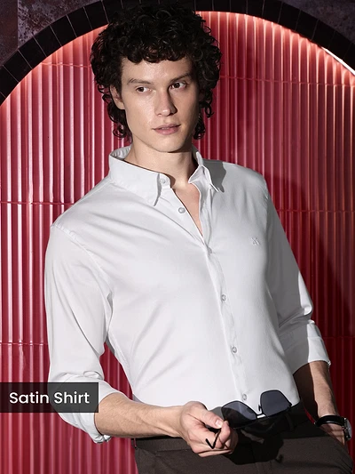 Man in a white satin shirts holding sunglasses, posing against a red background.